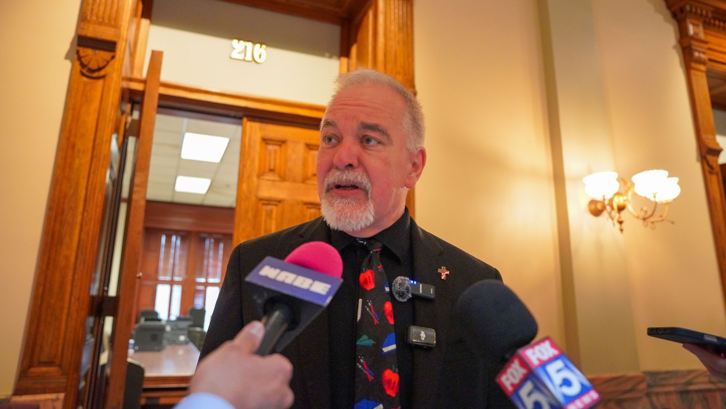 A man with a white beard and mustache talks to microphones in a hallway.