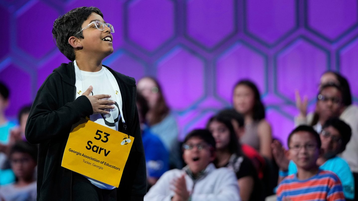 A boy competes in a spelling bee while standing in front of a crowd of other spellers