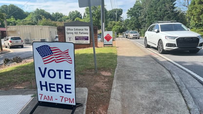 A large VOTE HERE sign on the side of a road, where multiple cars are parked