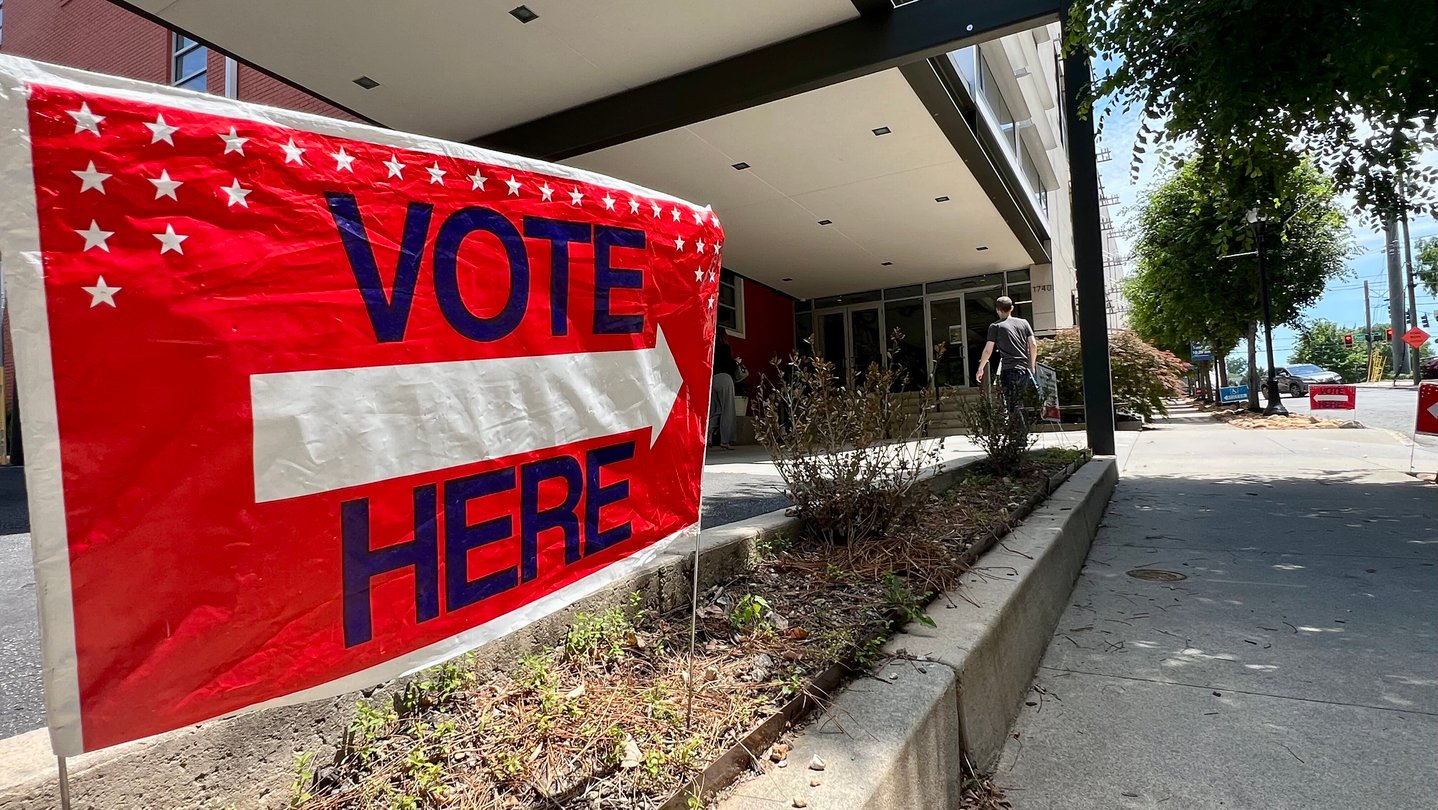 A vote here sign is shown in front of a church as a person in the background walks into the building.