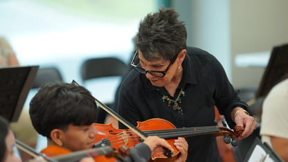 A woman stands, bending over with her hand on the scroll of a violin while a student plays it.