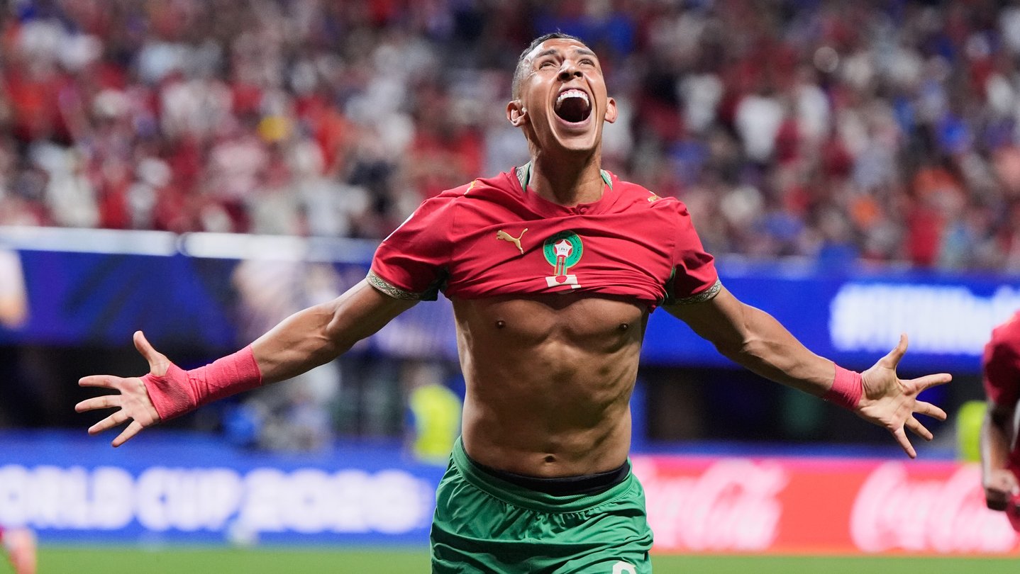 A soccer player wearing a red jersey holds his arms out in celebration