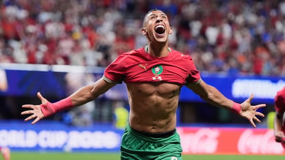 A soccer player wearing a red jersey holds his arms out in celebration