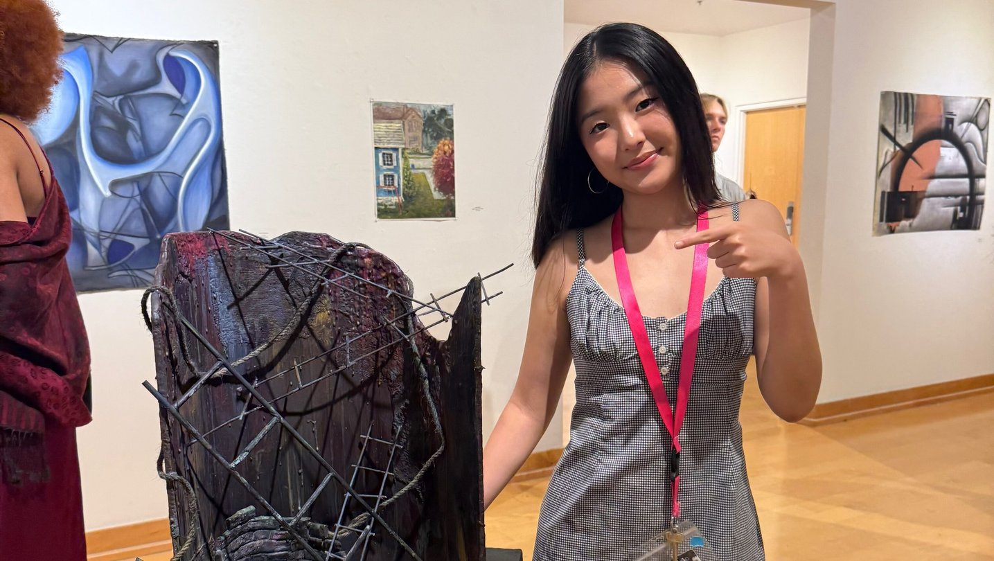 A teenage girl with long dark hair in a gray checkered dress stands next to a dark, textured mixed-media sculpture on a black pedestal in a gallery. She points at the work and smiles.
