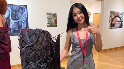 A teenage girl with long dark hair in a gray checkered dress stands next to a dark, textured mixed-media sculpture on a black pedestal in a gallery. She points at the work and smiles.