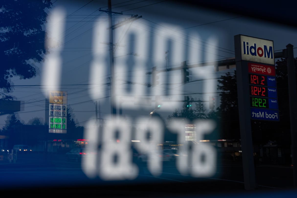 A close-up of a gas pump shows a price over 100 dollars, with a reflection of gas station signs in it