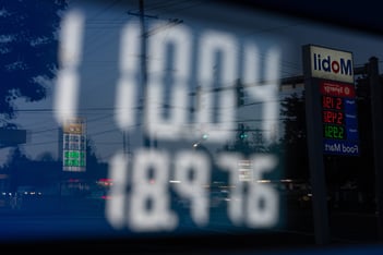 A close-up of a gas pump shows a price over 100 dollars, with a reflection of gas station signs in it