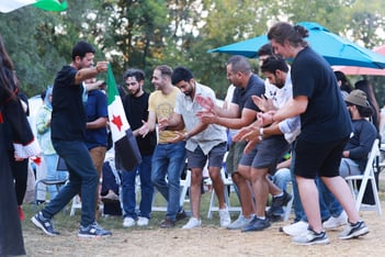 Men in matching headscarves dance dabkeh in a line, holding hands, at an outdoor event.