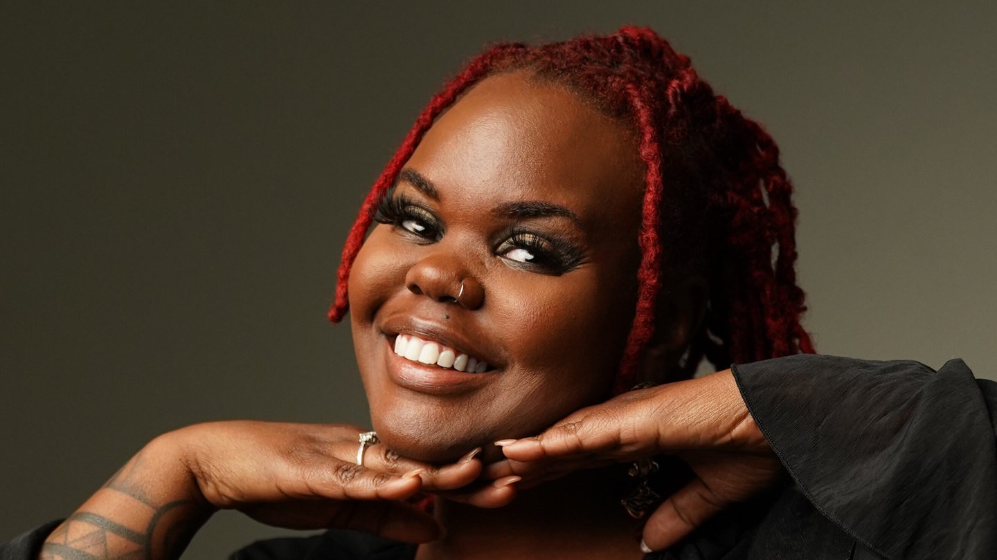 Ashlee Haze, wearing a black ruffled top and red locs, smiles with her chin resting on her hands against a dark studio backdrop.