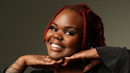 Ashlee Haze, wearing a black ruffled top and red locs, smiles with her chin resting on her hands against a dark studio backdrop.