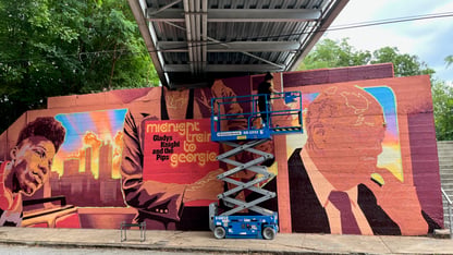 A muralist on a lift paints a portrait beside a colorful “Midnight Train to Georgia” mural panel in English Avenue.