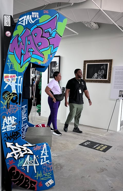 Two visitors walk past framed photography near the graffiti-covered WABE phone booth inside the Atlanta Cultural Exchange gallery space.