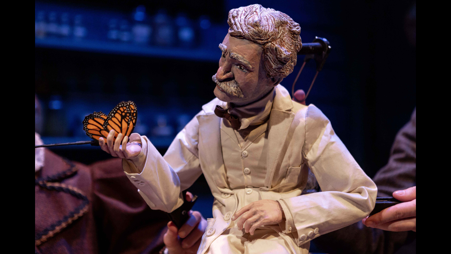 A puppet of an older man in a lab coat holds a monarch butterfly on stage in TESLA VS. EDISON.
