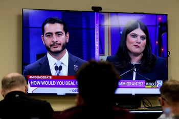 People watch a TV screen showing a political debate, with a main in a suit and tie on the left and a woman in a dress on the right