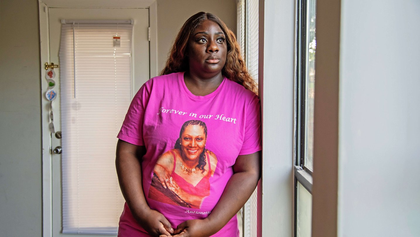 A woman in a pink t-shirts stands looking pensively out a window