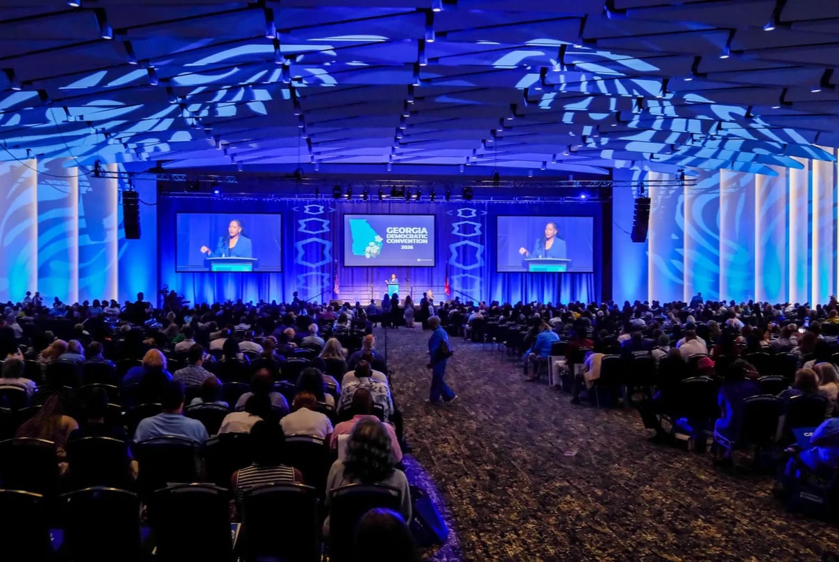 Hundreds of people sit in chairs in a large convention hall as a woman speaks on a stage bathed in blue lights