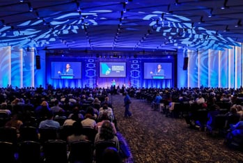 Hundreds of people sit in chairs in a large convention hall as a woman speaks on a stage bathed in blue lights
