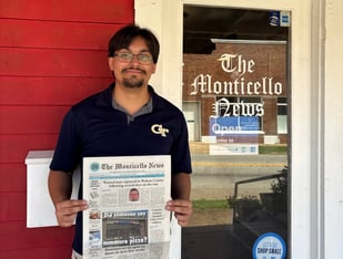 A man in a polo shirt stands holding a newspaper showing the front page next to a door that says the monticello news