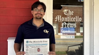 A man in a polo shirt stands holding a newspaper next to a glass door that say sthe monticello news