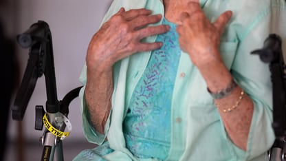 An elderly woman sits in a chair holding her hands to her chest