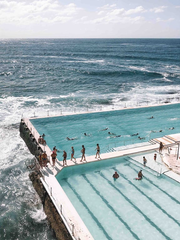 People entering and exiting the ocean to go to a pool that is in the middle of the ocean. | Panoramic view of the Blue Mountains in Australia. | Two people walking by black and white buildings. | Art gallery with different colored vases and paintings. | Bright red drink in a glass garnished with a cucumber. | Martini with a green olive in it and a tin can with the name "Mar-tinny" on it. | Four pieces of shellfish surrounding another piece of shellfish on a small bowl of tiny ice pieces. | Various types of pastry lined up in neat rows on a piece of parchment-like paper. | Hotel room with a bed, night table, and green plants. There is a large window that leads into another room. | Inside of a hotel. There is a brick wall in the back. In the room there are different chairs and couches that people can relax in.