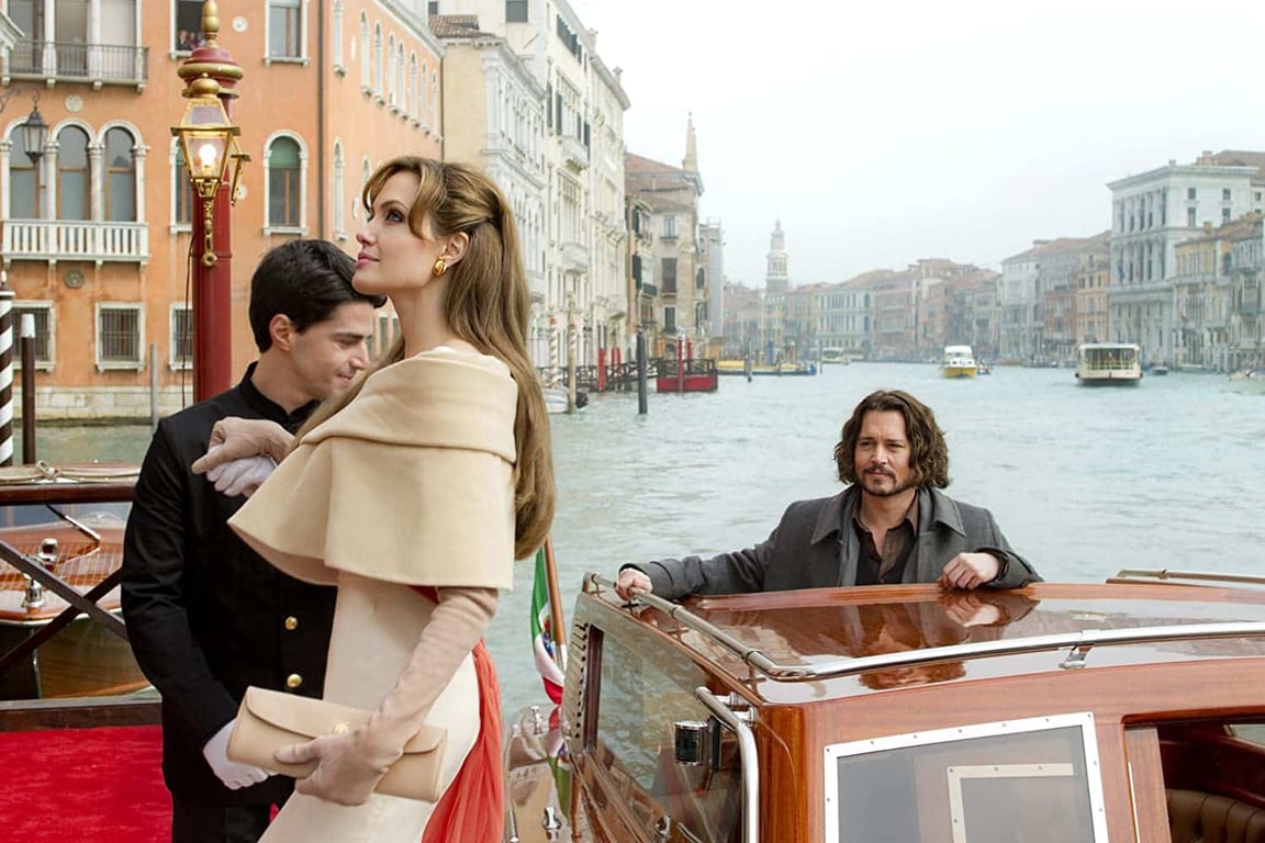 Scene from the movie The Tourist. Johnny Depp is on a boat in Italy with Angelina Jolie stepping onto a dock. | Scene from The Talented Mr. Ripley with Matt Damon, Jude Law, and Gwyneth Paltrow are sitting on a rock wall. | Scene from the movie Crazy Rich Asians with Henry Golding sitting by the water and showing his friend a ring. | Scene from the movie Midnight in Paris where a man and woman are walking down a cobblestone street. | Scene from The Darjeeling Limited movie where three men are carrying their luggage across a grassy field. | Scene from the movie Wild where Reese Witherspoon is carrying a large hiking pack and overlooking a body of water. | Scene from the movie Roman Holiday where a man and a woman are ready to clink their wine glasses with each other. | Scene from the movie Lost in Translation where a woman is looking out her window which overlooks a city skyline.
