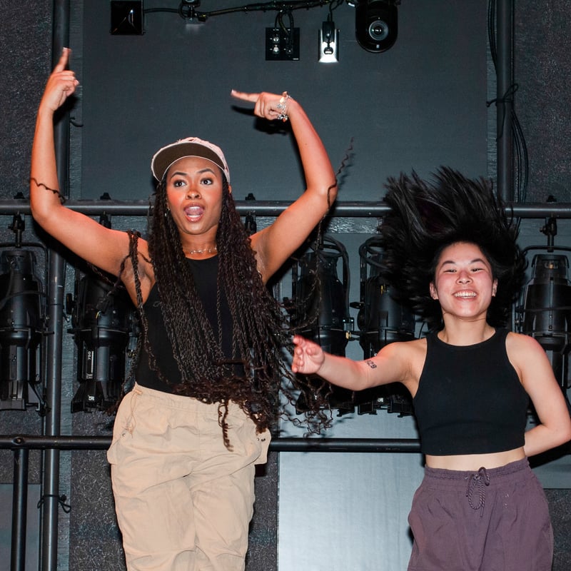 Two women jumping in a dance studio, expressing joy and energy, with dramatic lighting equipment in the background.