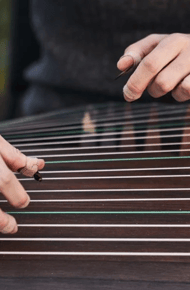 Close-up of hands playing a string instrument, with fingers positioned on the strings and a wooden surface visible.