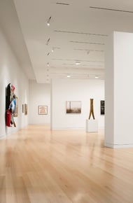 A spacious art gallery with white walls, wooden floors, and various framed artworks displayed. Bright lighting throughout.