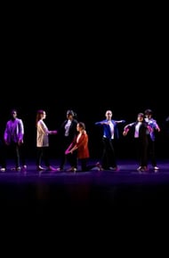 A group of dancers performing on stage under dim lighting, showcasing synchronized movements in colorful outfits.
