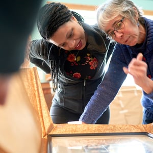 Two women examine a framed picture together, engaged in conversation, with a person’s hands visible in the foreground.