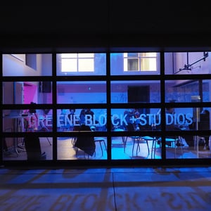 A dimly lit studio with people visible through large windows, featuring blue lighting and graffiti on the walls.