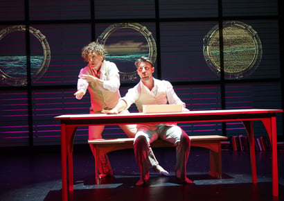 two men on stage at a red table looking to the side