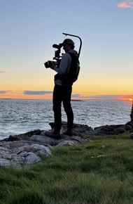 A silhouette of a photographer with a camera on a rocky shoreline at sunset, overlooking the ocean and a stone structure.