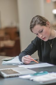 A student studying at a table, focusing on notes with a laptop and papers scattered around, engaged in discussion.