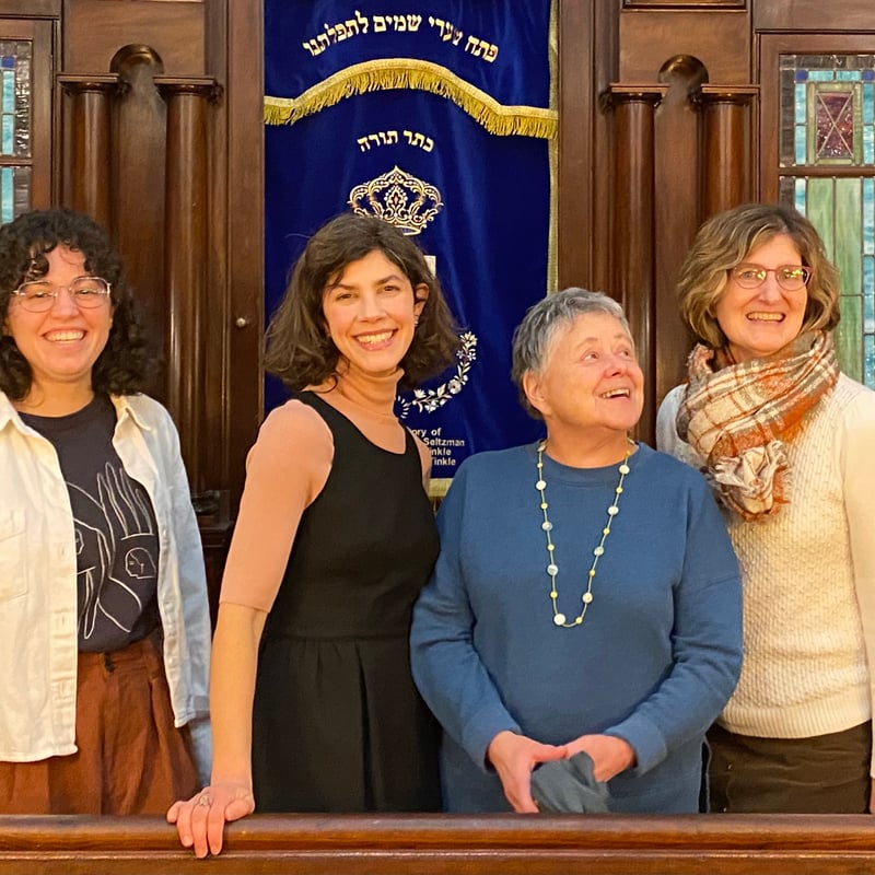 A group of six smiling women stands together in front of a decorative blue banner in a warmly lit interior