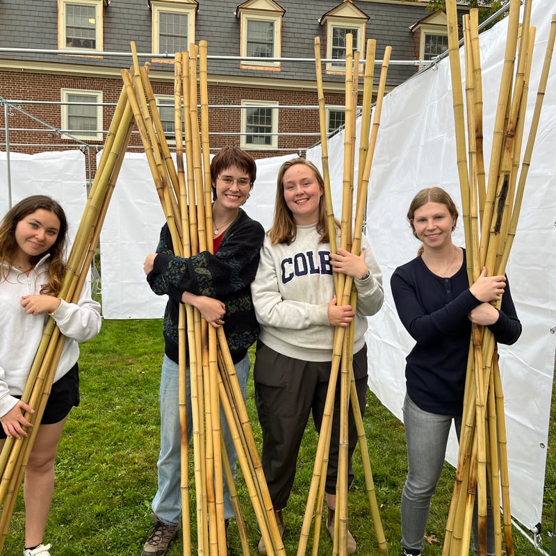 Four young women are smiling while holding bundles of bamboo poles outdoors with a tent structure in the background