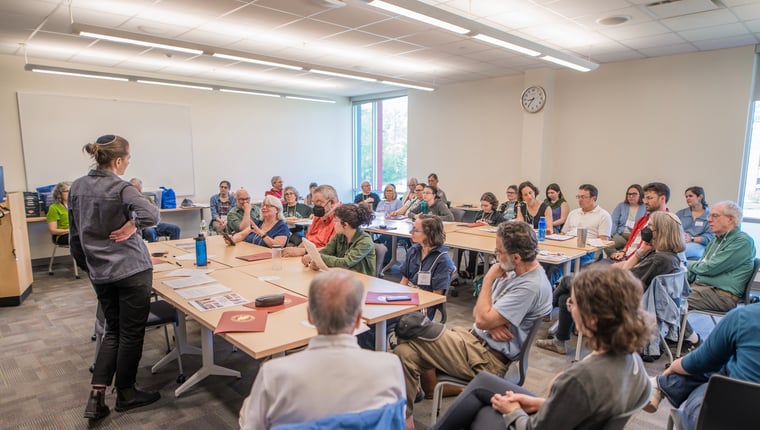 A group of people attentively watching a speaker in a classroom setting with desks and a whiteboard