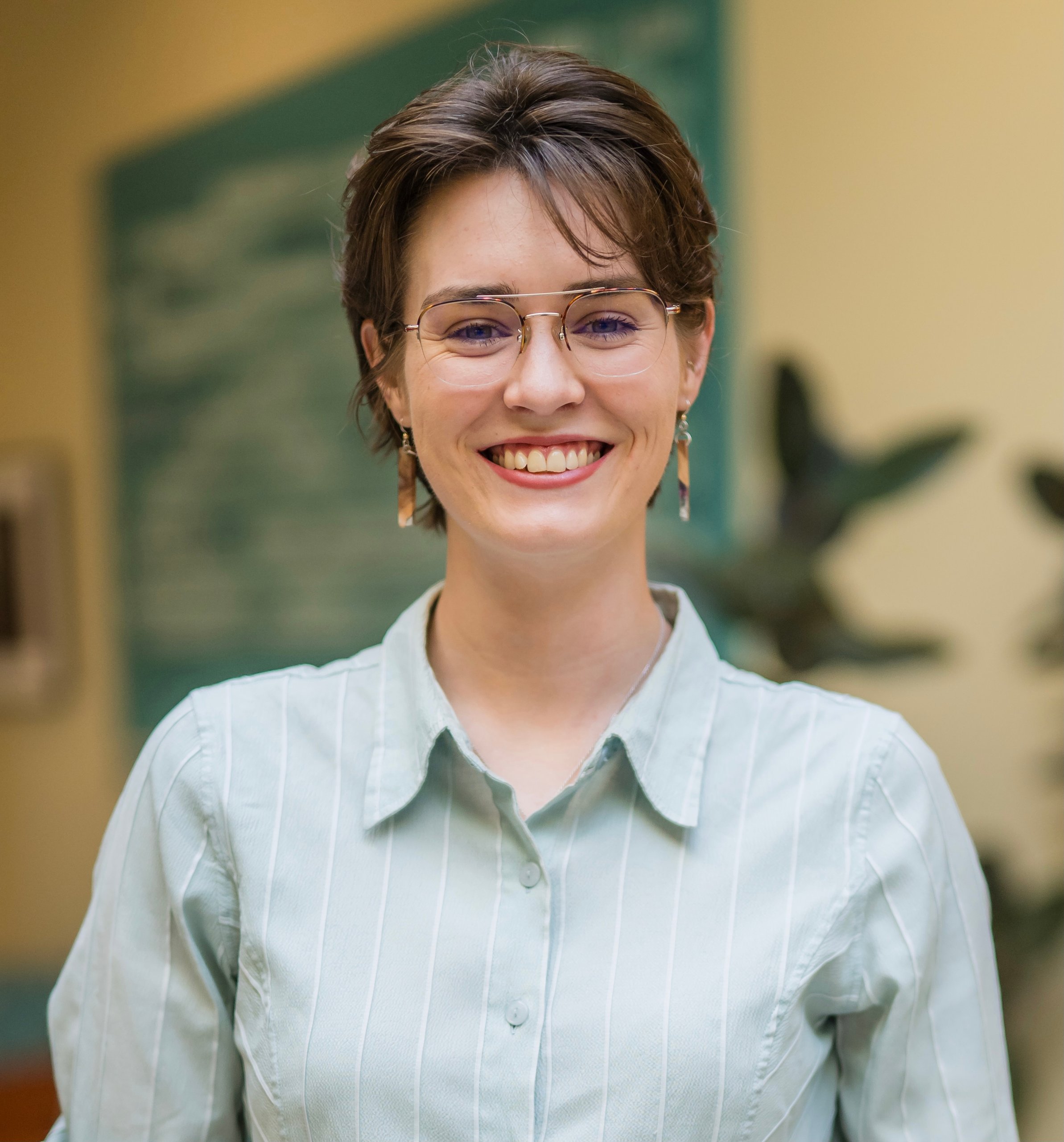 Smiling young woman with short hair glasses and a light green shirt standing indoors with plants in the background