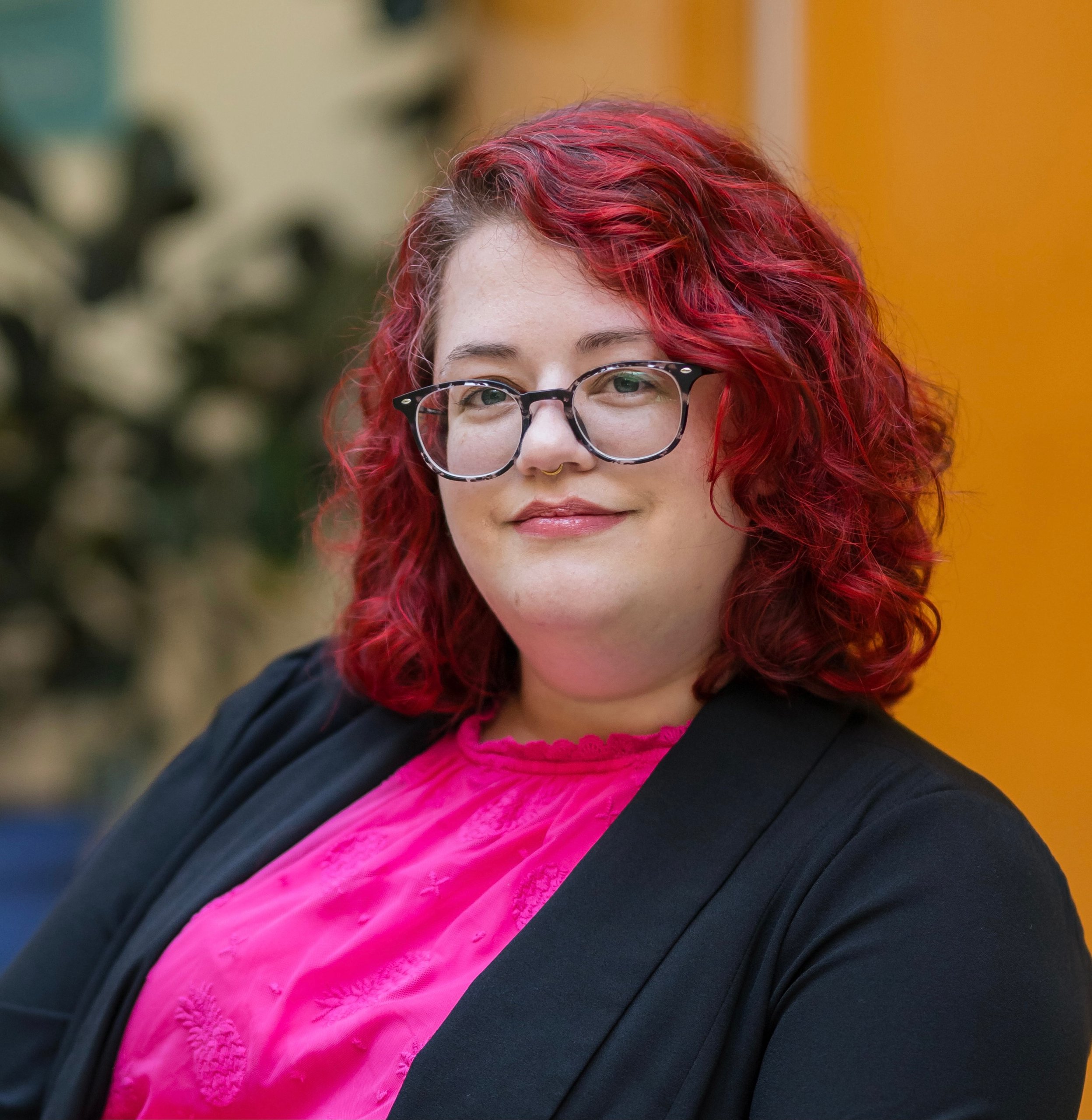 Person with curly red hair and glasses wearing a pink top and black cardigan smiling in a welllit indoor setting