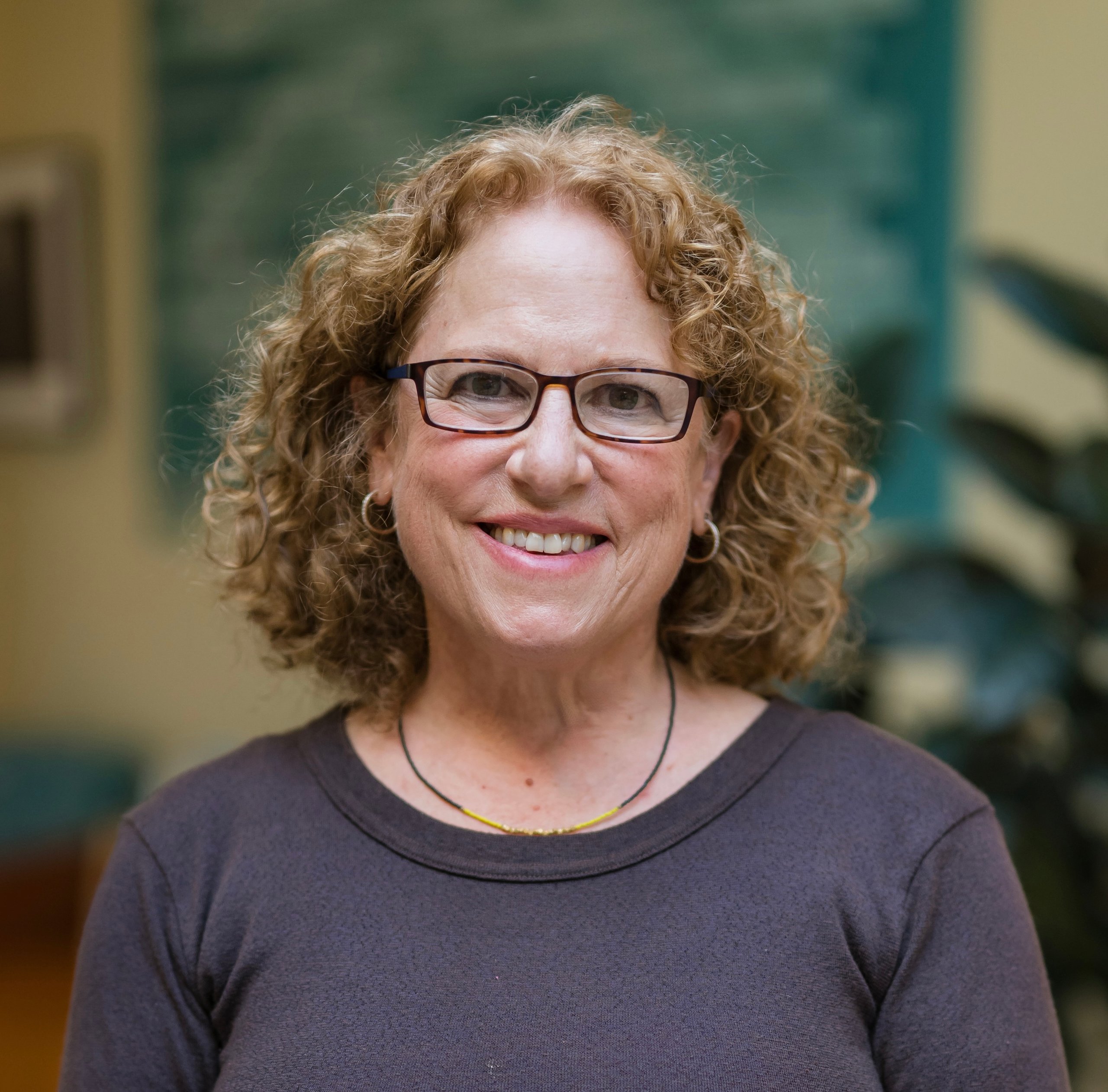 Woman with curly hair and glasses smiling wearing a dark sweater with indoor plants and a soft background in view