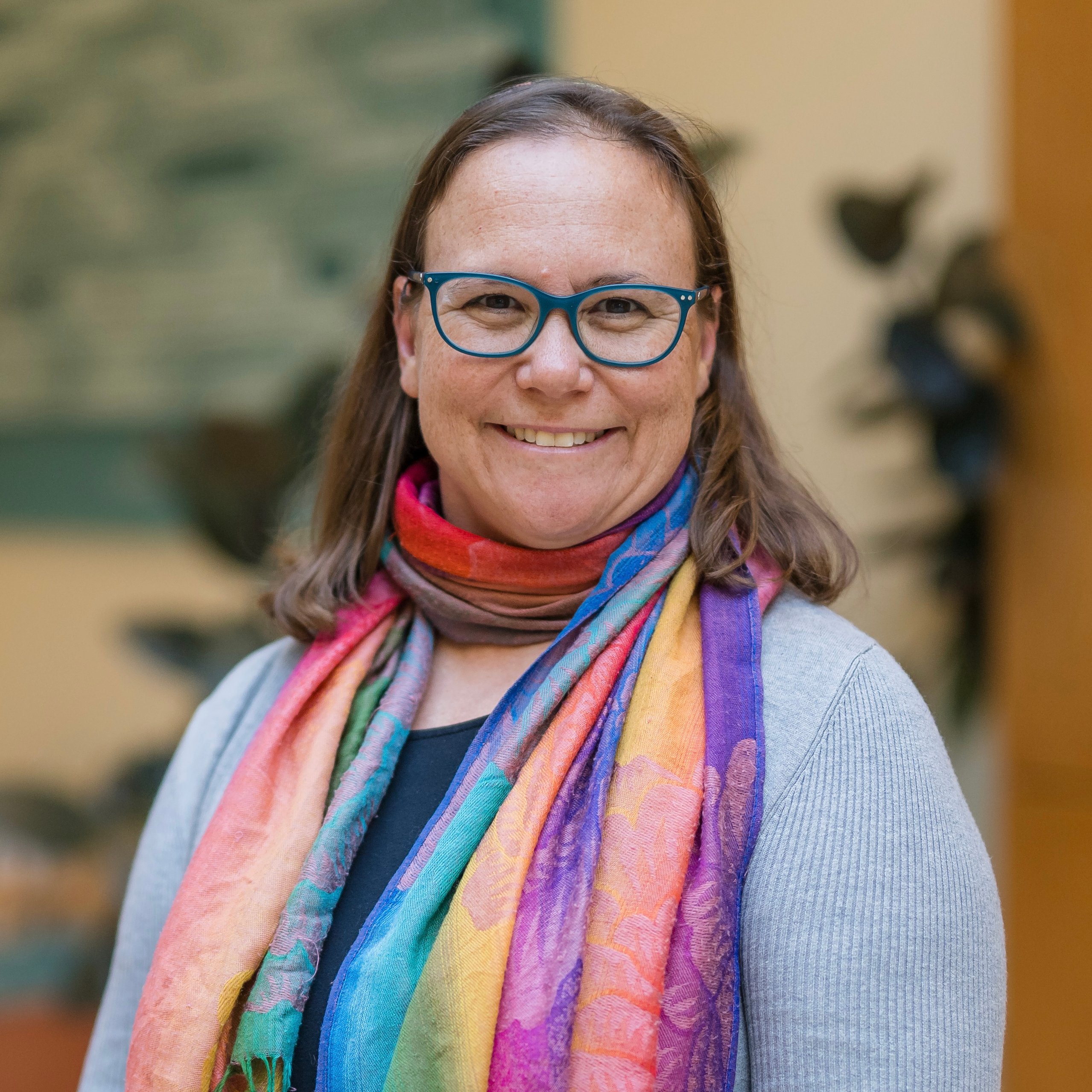 Smiling woman with glasses wearing a colorful scarf and a gray sweater standing in a bright indoor setting