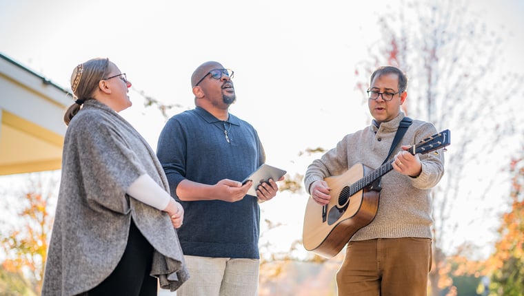 Three people outdoors singing together one playing guitar with a warm autumn backdrop and soft sunlight