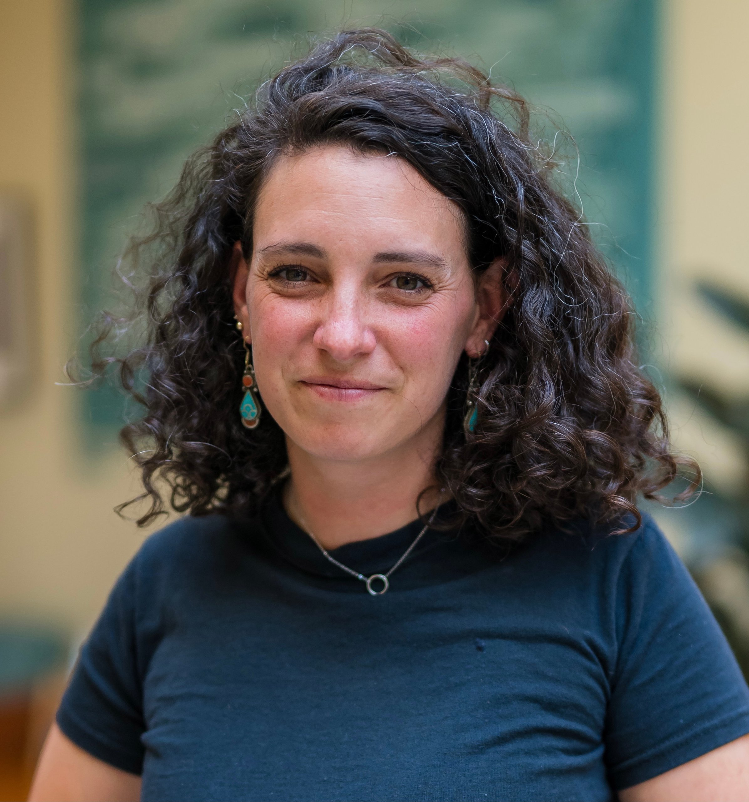 A woman with curly hair wearing a black shirt and earrings smiling softly in a bright indoor setting
