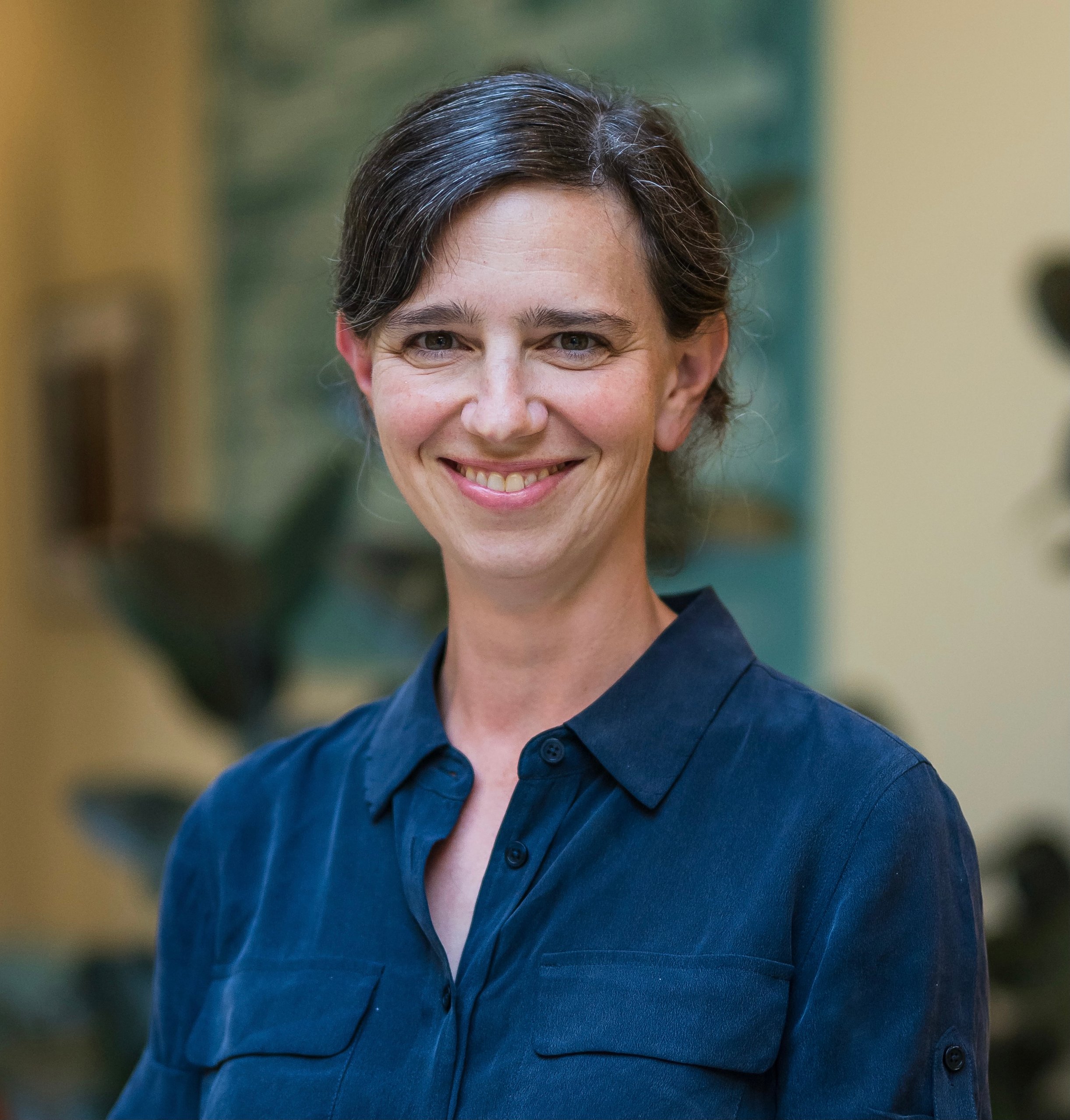 Portrait of a woman with shoulderlength hair smiling wearing a blue shirt in a softly lit interior setting with plants