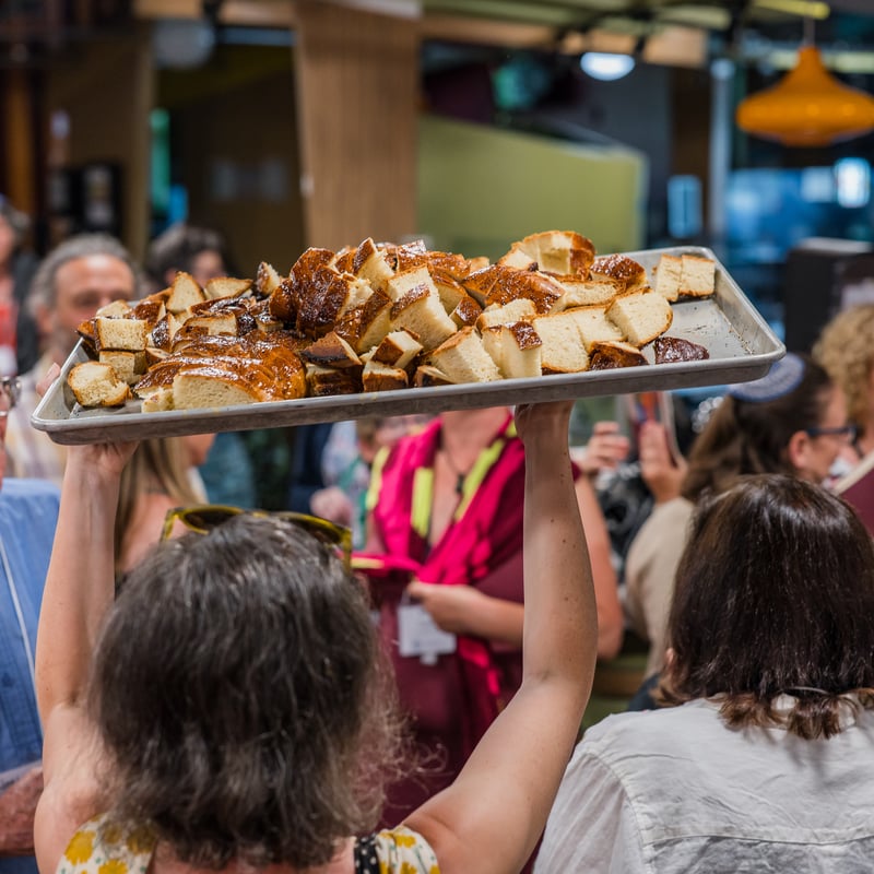 A person holds a tray of bread above a crowd in a lively market setting with people chatting and enjoying the atmosphere