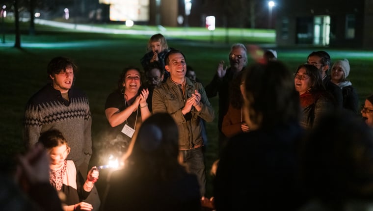 Group of people gathered around a fire at night smiling and clapping enjoying a lively outdoor gathering