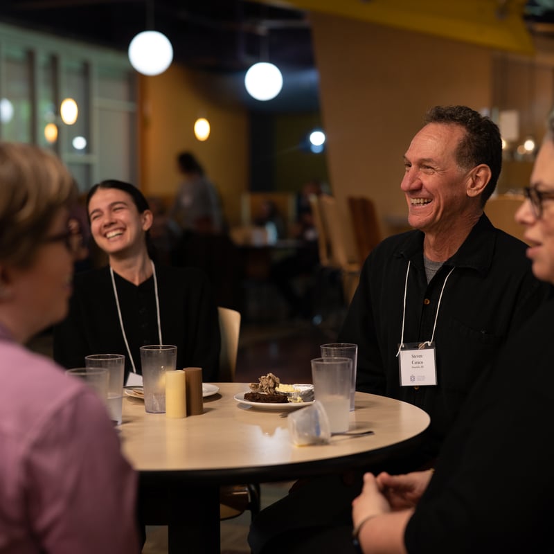 Group of people laughing and chatting around a table in a cozy setting with drinks and dessert visible