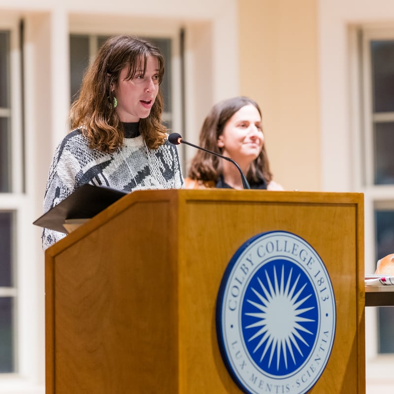 Two women speak at a podium with a Colby College logo while a man sits in the background A table has bread