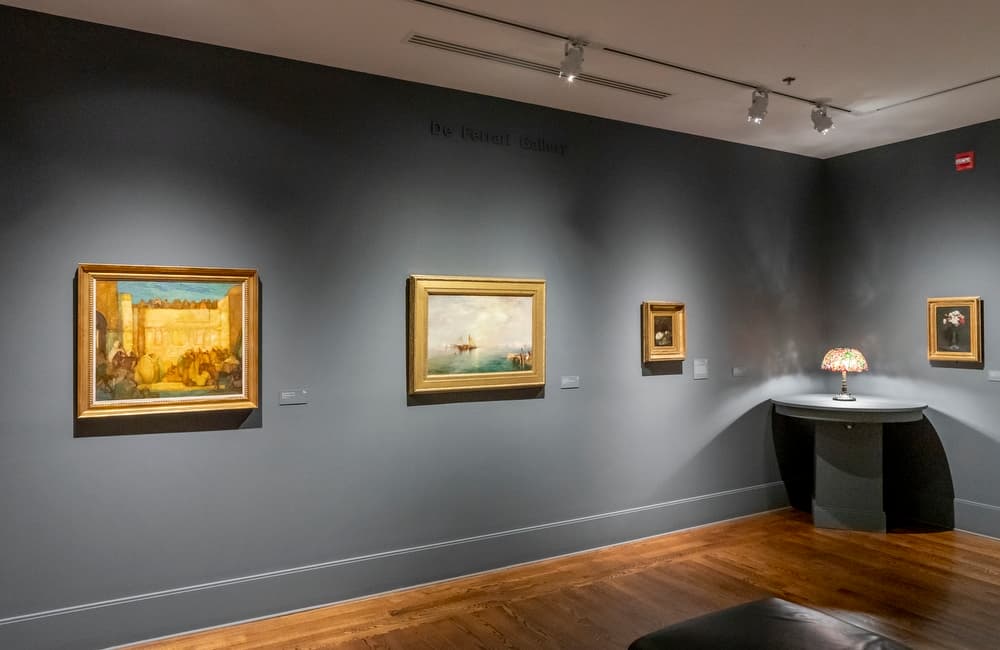 A stained glass lamp and four paintings in gold frames hung in a gallery with grey walls